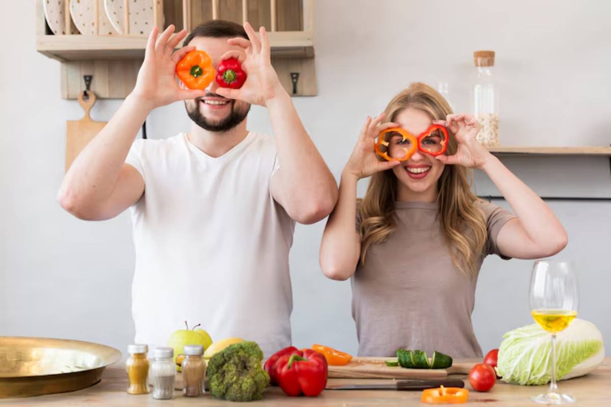 A man and woman hold orange bell peppers in front of their faces, surrounded by fresh vegetables and a glass of wine on a kitchen countertop.