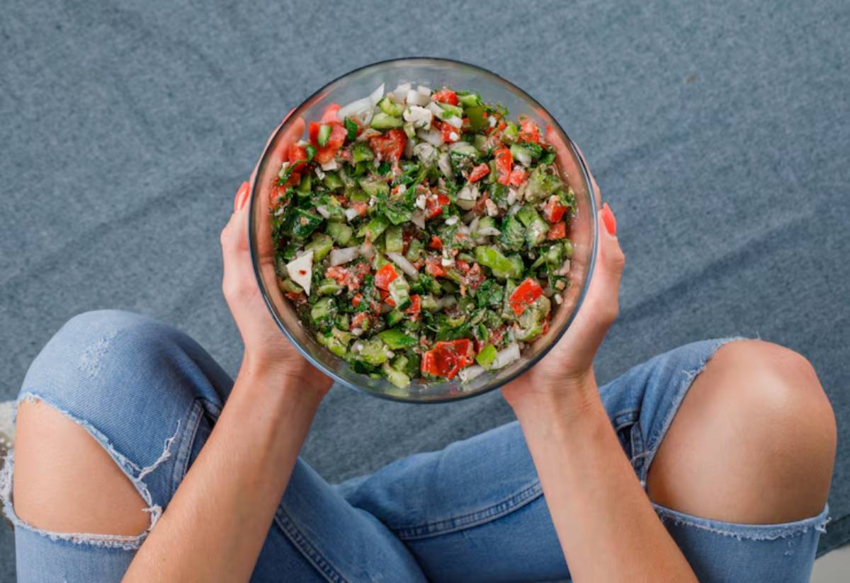 A person sitting cross-legged holds a large glass bowl filled with a colorful, fresh salad of herbs, tomatoes, and vegetables.
