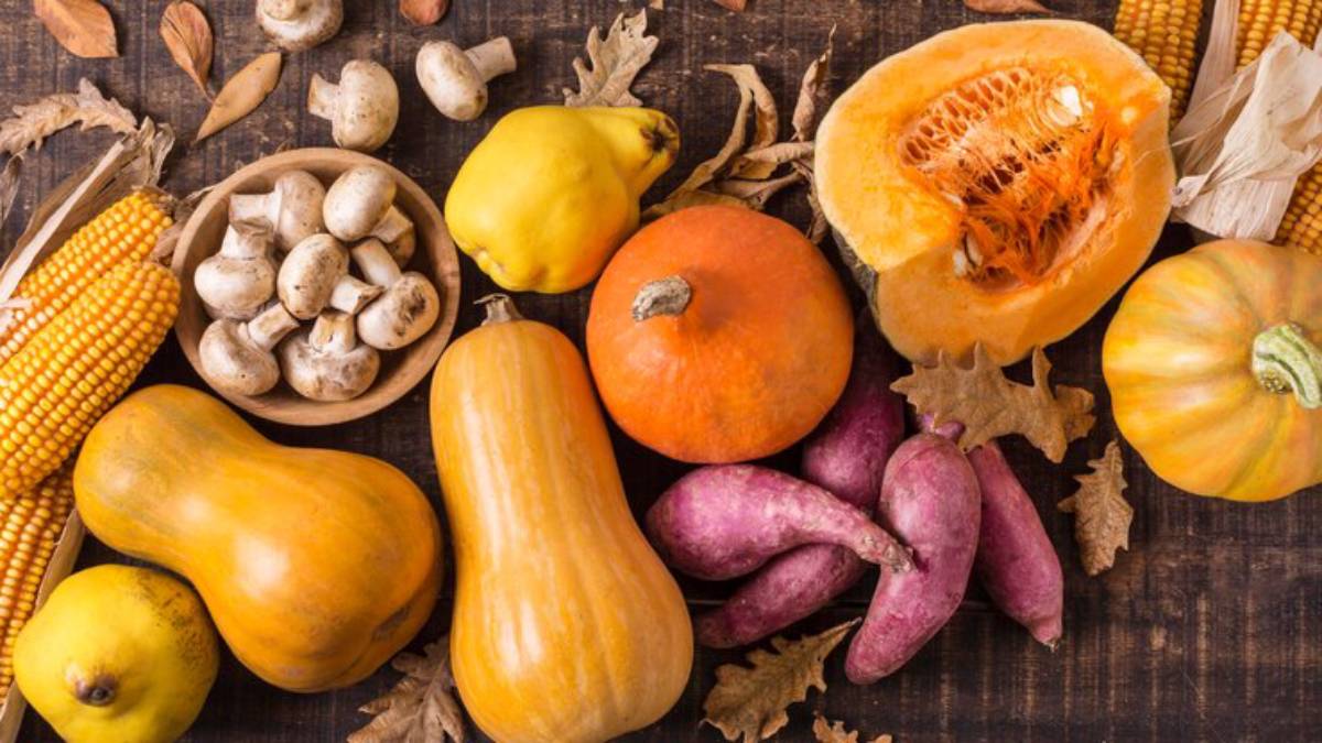 A colorful assortment of autumn produce, including pumpkins, squash, sweet potatoes, mushrooms, corn, and decorative leaves on wood.