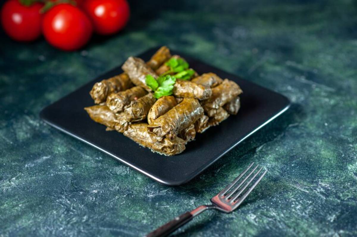 A black plate holds a stack of vine leaf wraps topped with cilantro, alongside a fork and red tomatoes in the background.