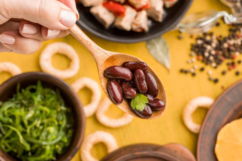 A hand holding a wooden spoon filled with kidney beans garnished with a sprig of cilantro, with various food items in the background.