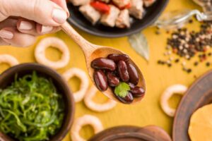A hand holding a wooden spoon filled with kidney beans garnished with a sprig of cilantro, with various food items in the background.