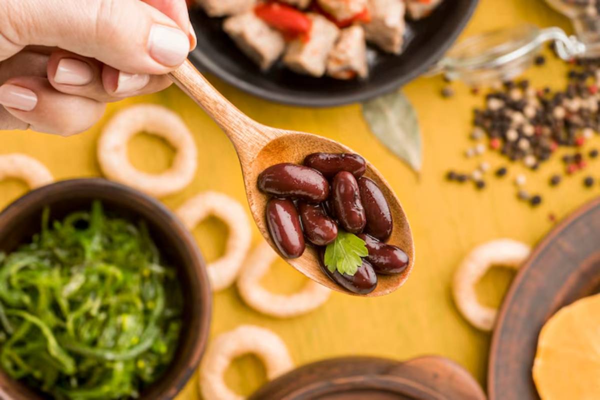 A hand holding a wooden spoon filled with kidney beans garnished with a sprig of cilantro, with various food items in the background.