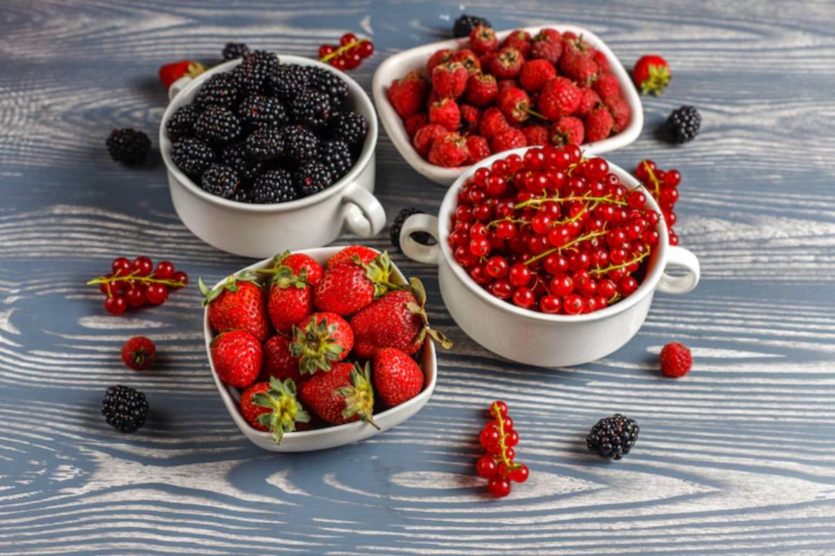 Four bowls filled with fresh berries: blackberries, strawberries, raspberries, and red currants, arranged on a wooden surface.