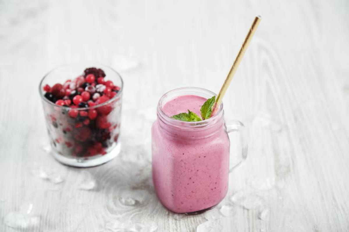 A creamy pink smoothie with a mint leaf garnish in a glass mug, alongside a cup of mixed frozen berries on a wooden surface.