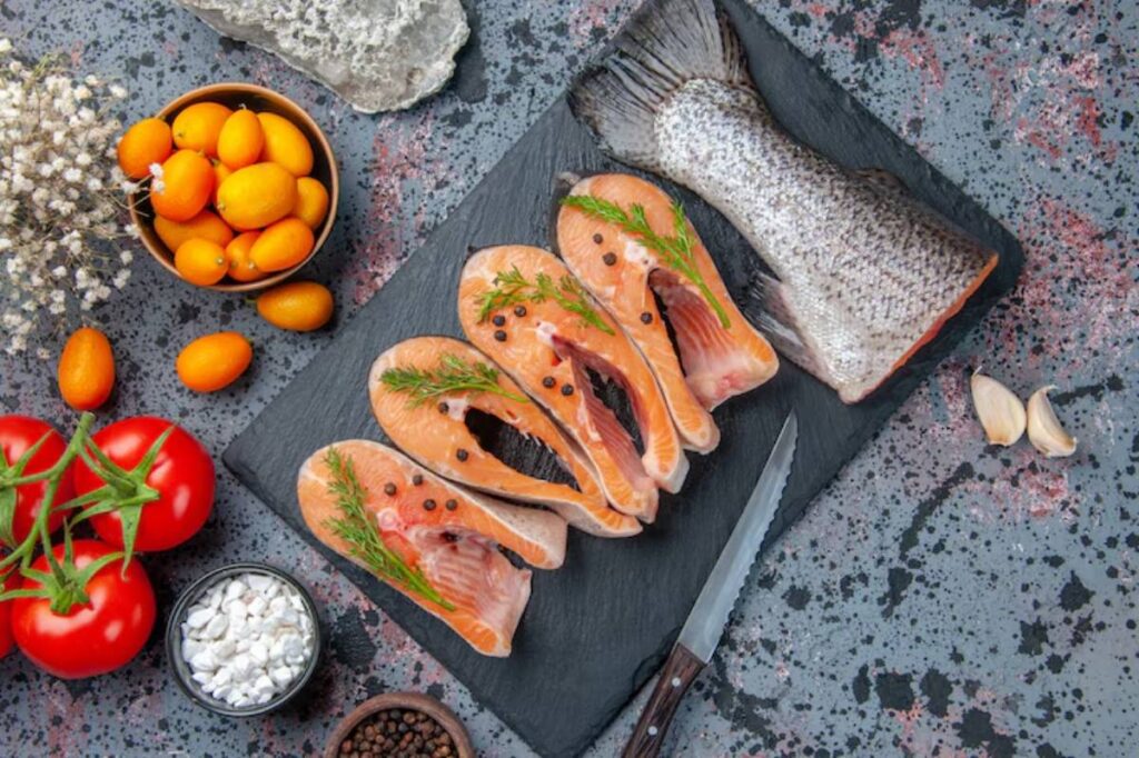A slate board displays salmon fillets with herbs, tomatoes, kumquats, garlic, and spices, creating a vibrant culinary scene.