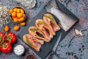 A slate board displays salmon fillets with herbs, tomatoes, kumquats, garlic, and spices, creating a vibrant culinary scene.