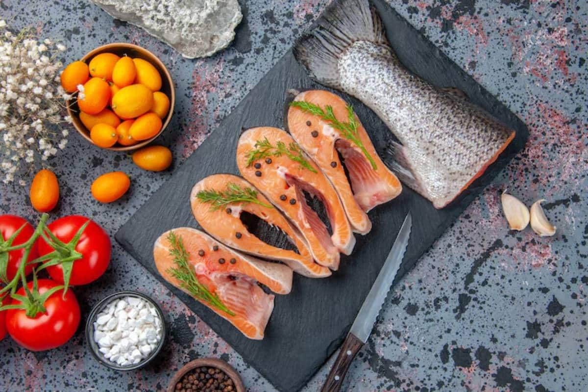 A slate board displays salmon fillets with herbs, tomatoes, kumquats, garlic, and spices, creating a vibrant culinary scene.