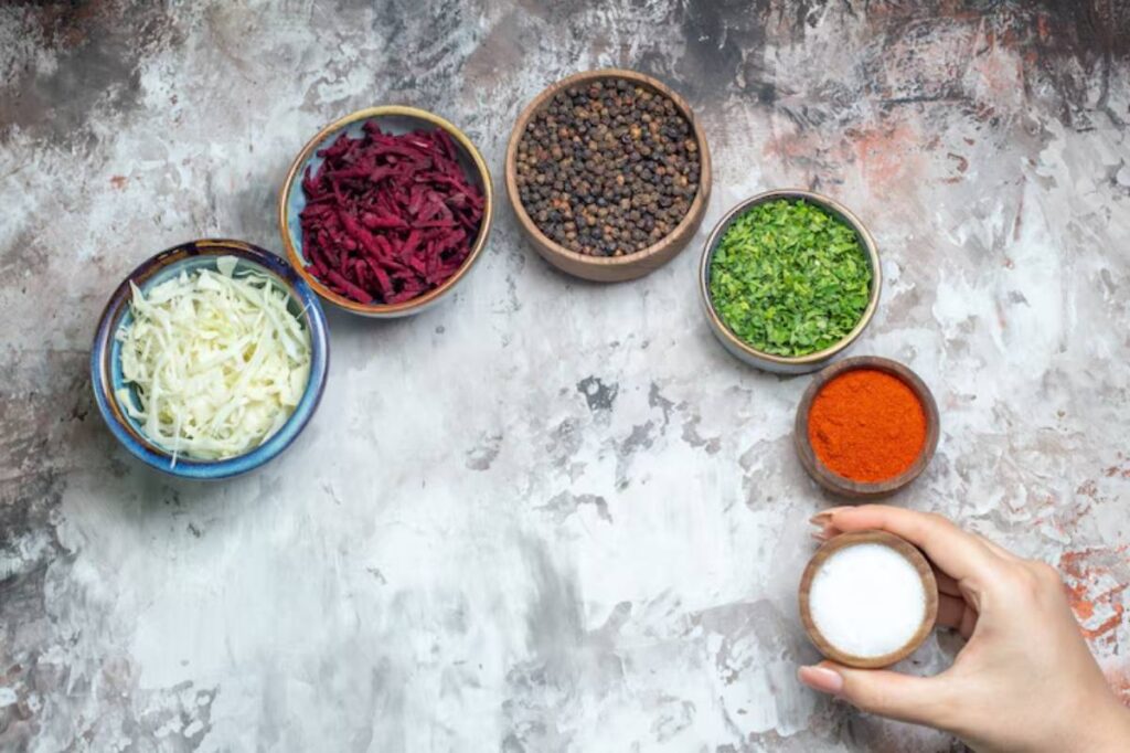 A hand holds a small bowl of salt next to vibrant bowls of shredded cabbage, beets, spices, and chopped herbs on a textured surface.