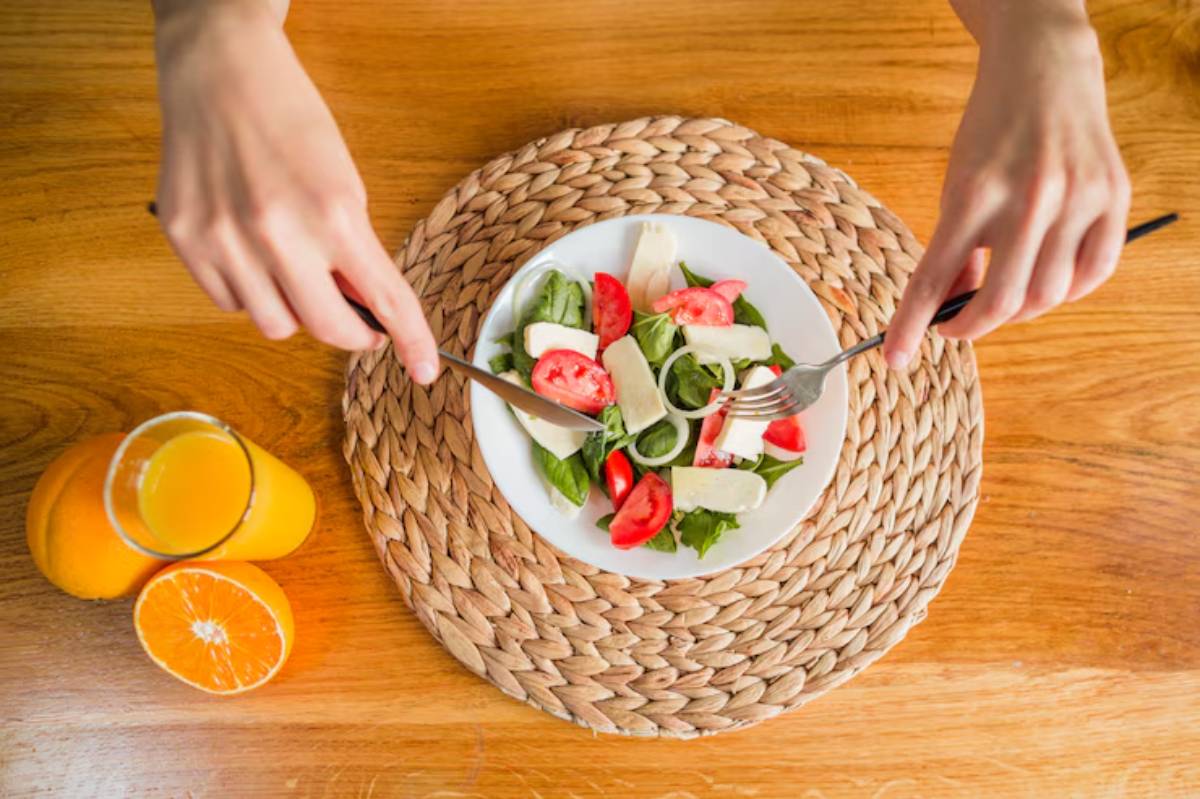 A close-up of hands holding forks over a fresh salad with tomatoes and cheese, beside a glass of orange juice and a halved orange.