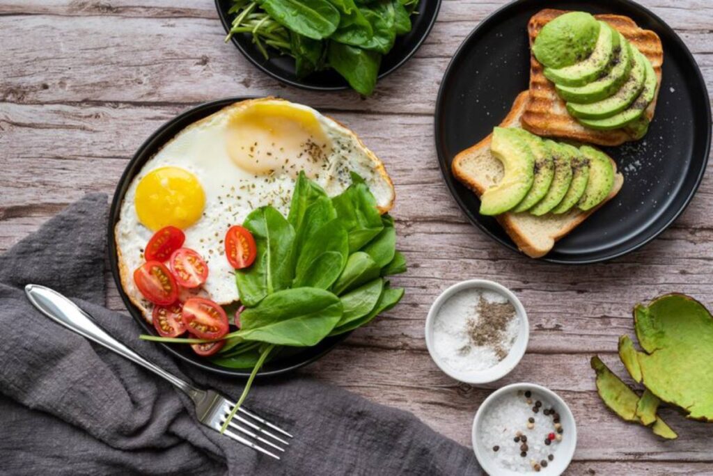 A healthy breakfast plate featuring fried eggs, cherry tomatoes, spinach, avocado toast, and seasonings on a wooden table.