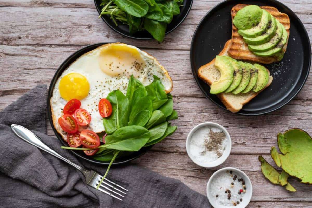 A healthy breakfast plate featuring fried eggs, cherry tomatoes, spinach, avocado toast, and seasonings on a wooden table.