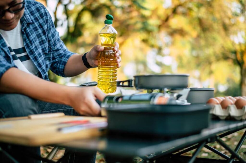 A person holds a bottle of cooking oil while preparing a meal outdoors, surrounded by cooking utensils and eggs on a table.