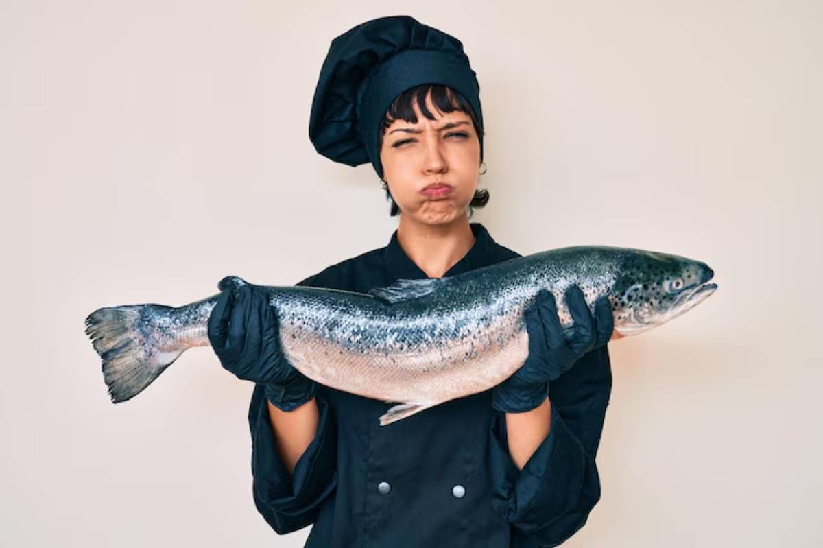 A chef wearing a black uniform and gloves holds a large, fresh salmon against a neutral background.