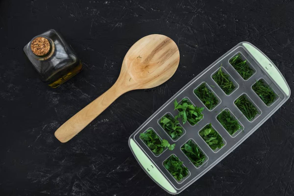 An olive oil bottle, wooden spoon, and an ice tray filled with chopped herbs on a dark background.