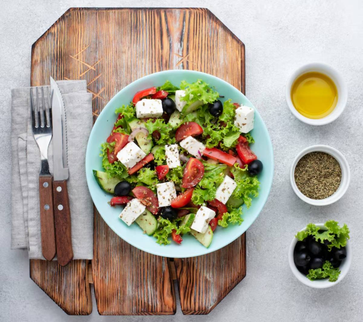 A fresh salad with lettuce, tomatoes, cucumbers, feta cheese, and olives, accompanied by olive oil and oregano in small bowls.
