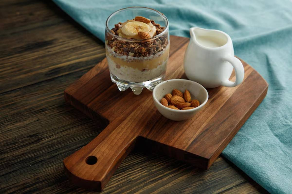 A glass dessert with layers of cream, granola, and banana, served on a wooden board with a bowl of almonds and a small milk pitcher.