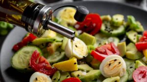 A sleek oil dispenser drizzles olive oil over a fresh salad with cucumbers, cherry tomatoes, boiled eggs, and avocado.