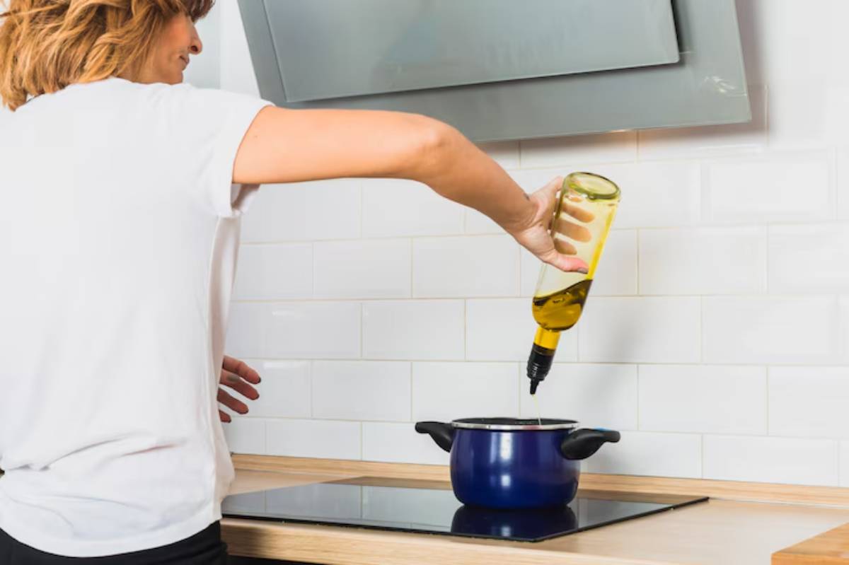 A person pours olive oil from a bottle into a blue pot on a modern stovetop, surrounded by a clean, bright kitchen.