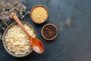 A bowl of cooked bulgur with a wooden spoon, accompanied by bowls of yellow grains and black pepper, on a dark stone surface.