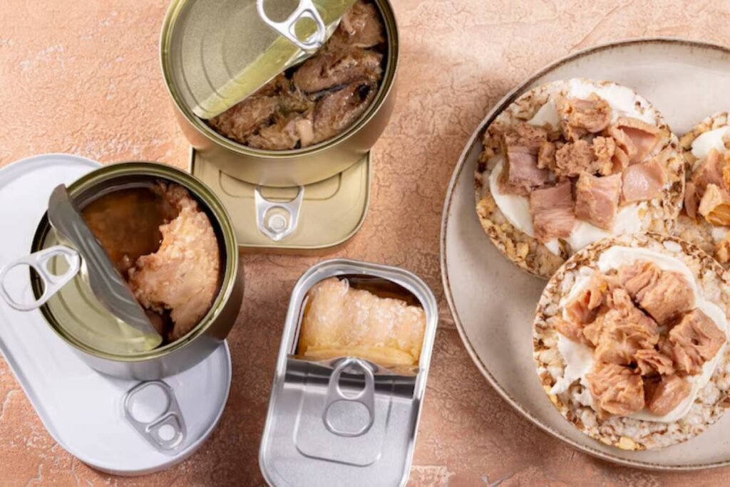 Three open cans of various meats beside rice cakes topped with chunks of tuna and chicken on a beige textured surface.