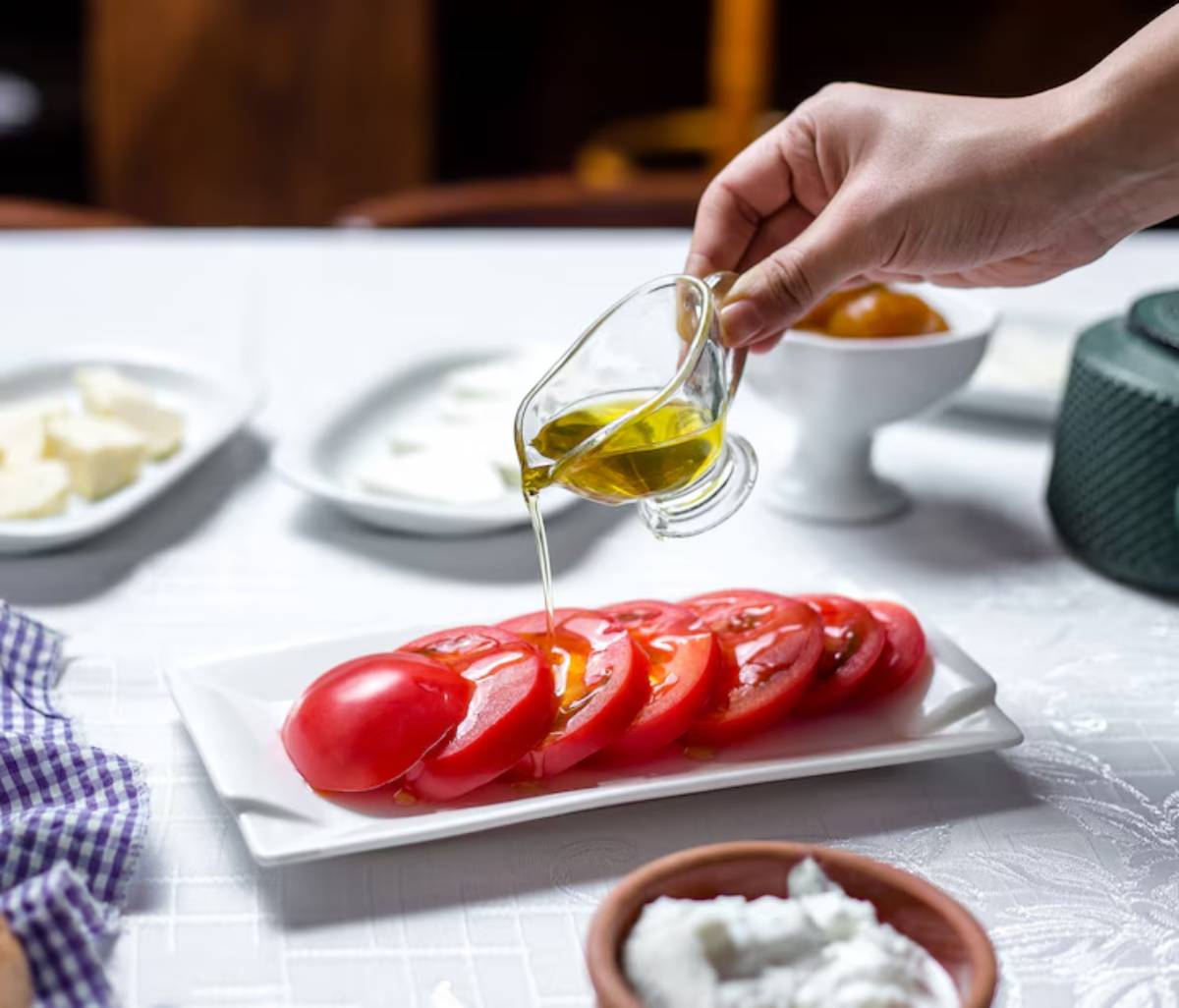 A hand drizzles olive oil over sliced tomatoes on a white plate, surrounded by various ingredients including cheese and a bowl of olives.
