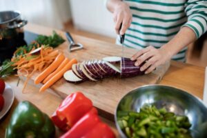 A person slices eggplant on a wooden cutting board, surrounded by carrots, peppers, and a pot in a bright kitchen.