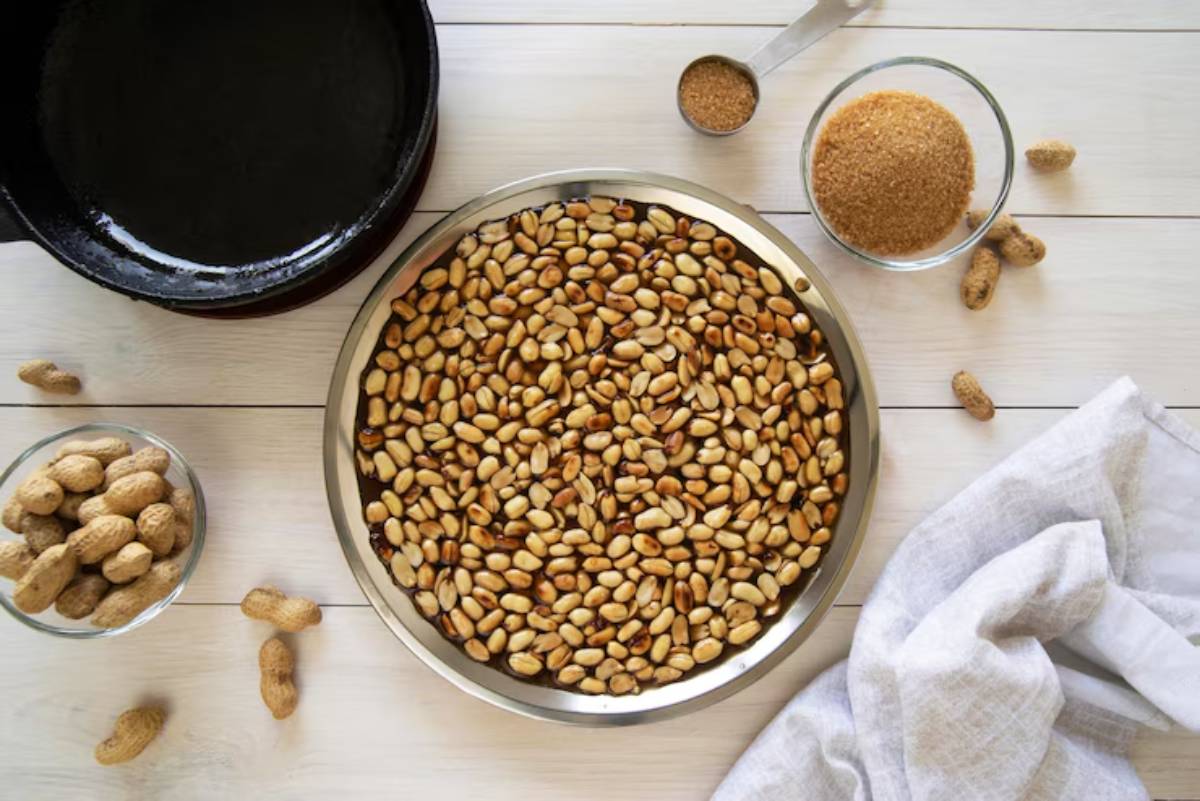 A tray of roasted peanuts surrounded by raw peanuts, sugar, and a frying pan on a wooden table.