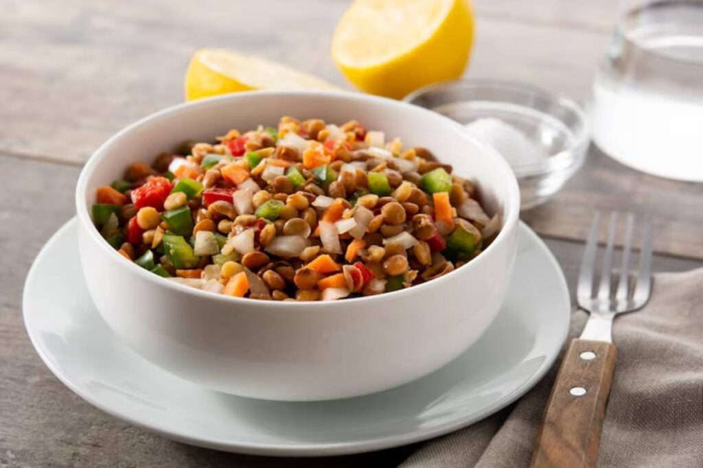 A white bowl filled with colorful lentil salad mixed with diced vegetables, lemon wedge, and seasonings on a wooden table.