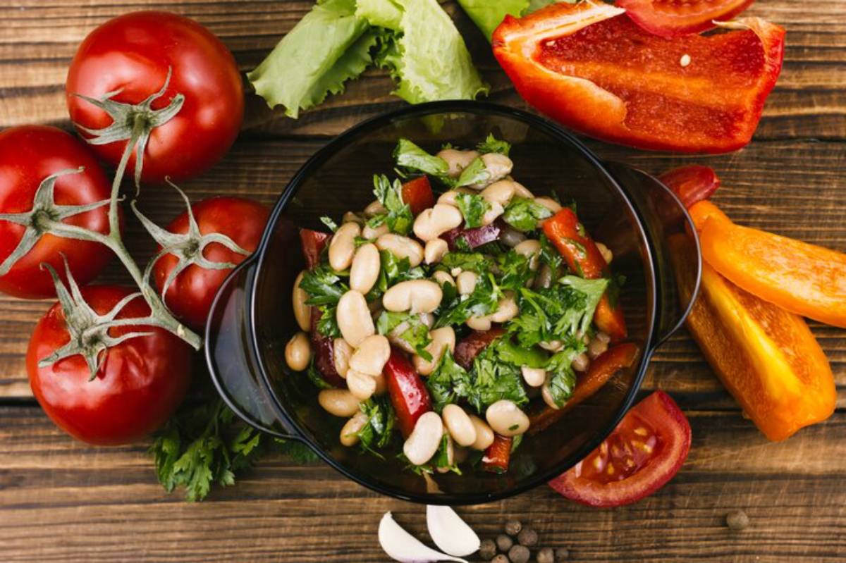 A vibrant salad of white beans, red and yellow peppers, and herbs, surrounded by fresh tomatoes and lettuce on a wooden surface.