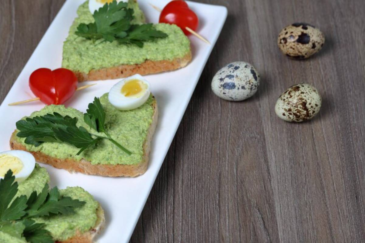 Heart-shaped toast topped with green spread, parsley, and quail eggs, accompanied by cherry tomatoes and decorative quail eggs.