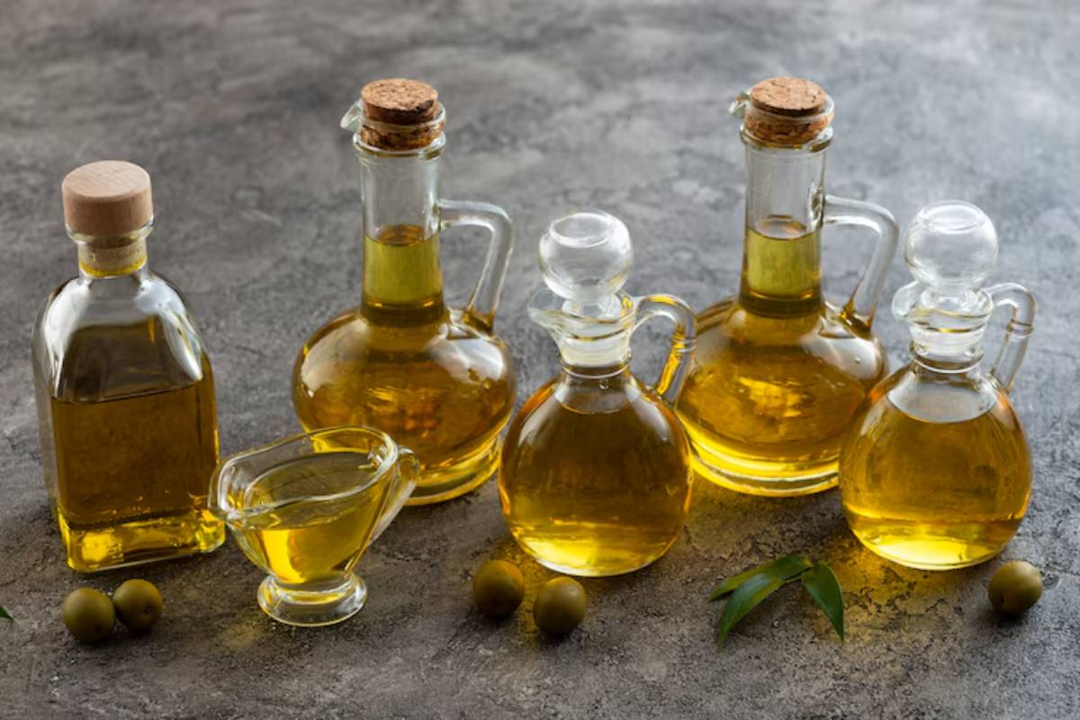 Five glass bottles filled with olive oil sit on a textured gray surface, accompanied by two green olives and a few leaves.