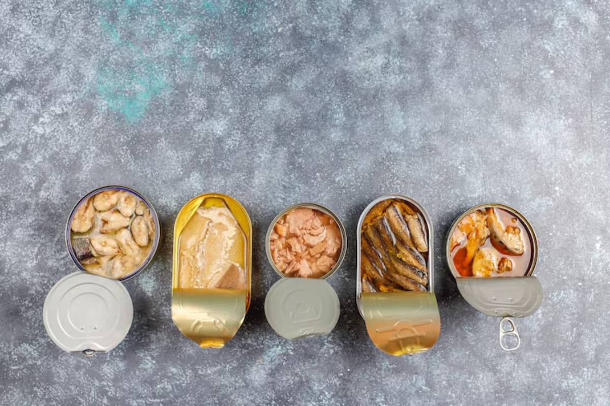 Five open cans displaying assorted seafood: shrimp, fish fillets, tuna, sardines, and canned meat, set on a textured gray surface.