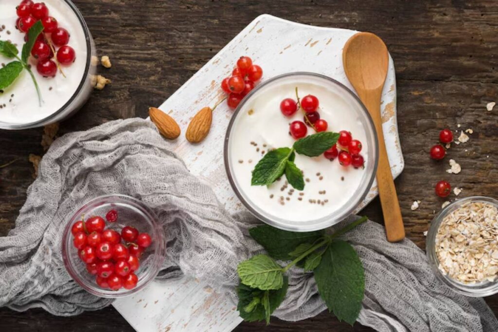A bowl of yogurt topped with red currants and mint, surrounded by almonds, oats, and a gray cloth on a rustic wooden surface.