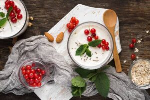A bowl of yogurt topped with red currants and mint, surrounded by almonds, oats, and a gray cloth on a rustic wooden surface.