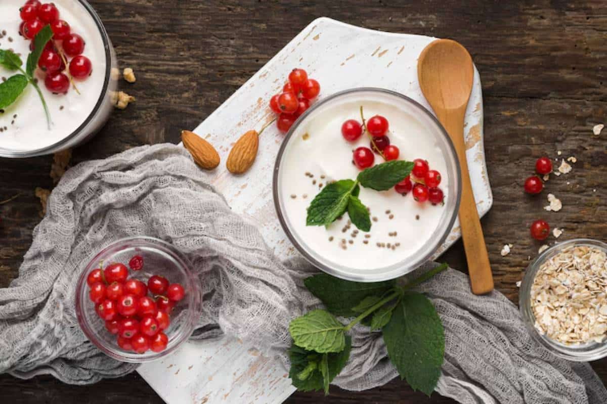 A bowl of yogurt topped with red currants and mint, surrounded by almonds, oats, and a gray cloth on a rustic wooden surface.