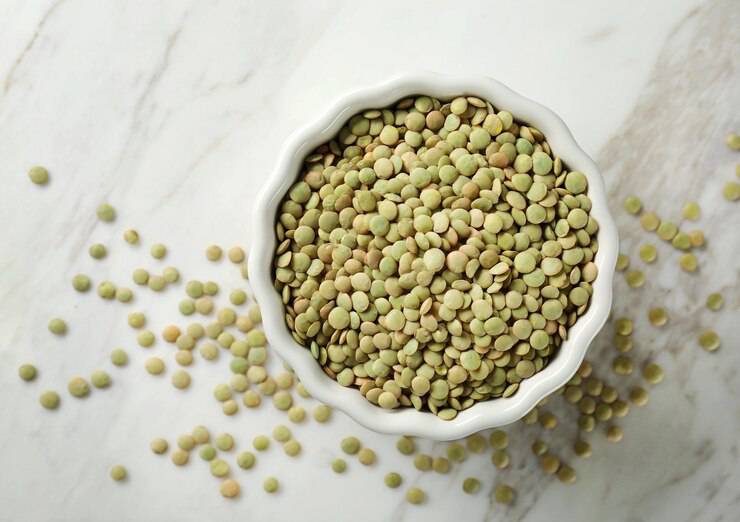 A white bowl filled with green lentils sits on a marble countertop, with some lentils scattered around.