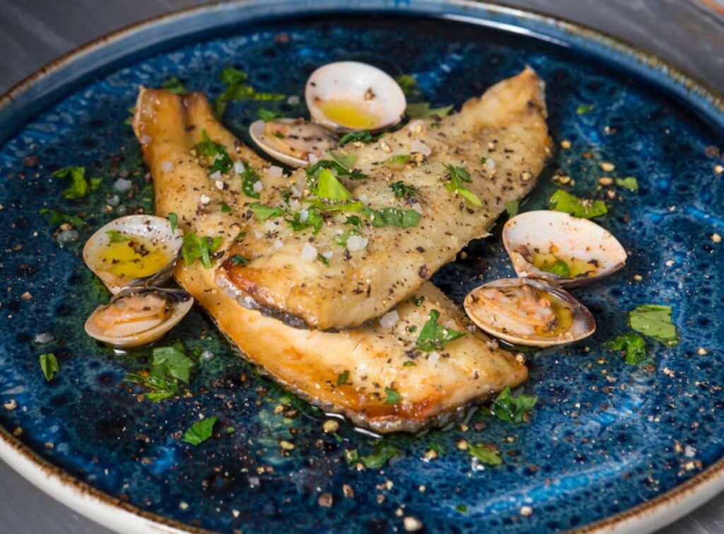A beautifully plated dish featuring pan-seared fish fillets garnished with clams, parsley, and olive oil on a blue ceramic plate.