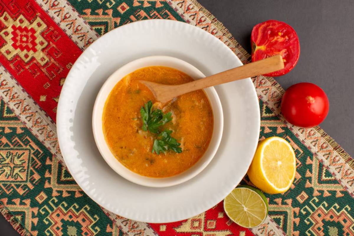 Bowl of flavorful soup garnished with fresh herbs, accompanied by tomatoes and citrus fruits, on a patterned fabric background.