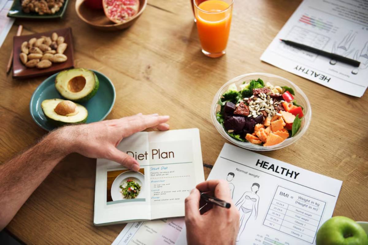 A person writes in a diet plan notebook surrounded by healthy foods, including a salad, fruits, nuts, and health charts.