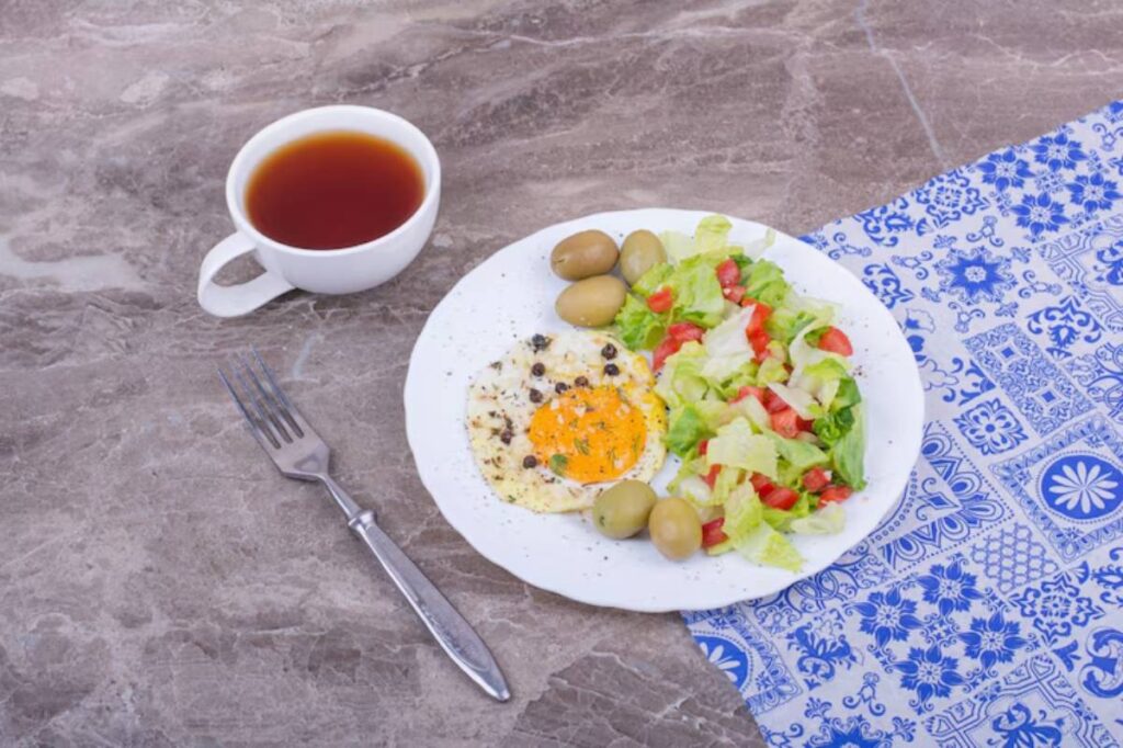 A plate with a sunny-side-up egg, salad, olives, and a cup of tea on a patterned tablecloth.
