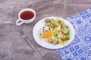A plate with a sunny-side-up egg, salad, olives, and a cup of tea on a patterned tablecloth.