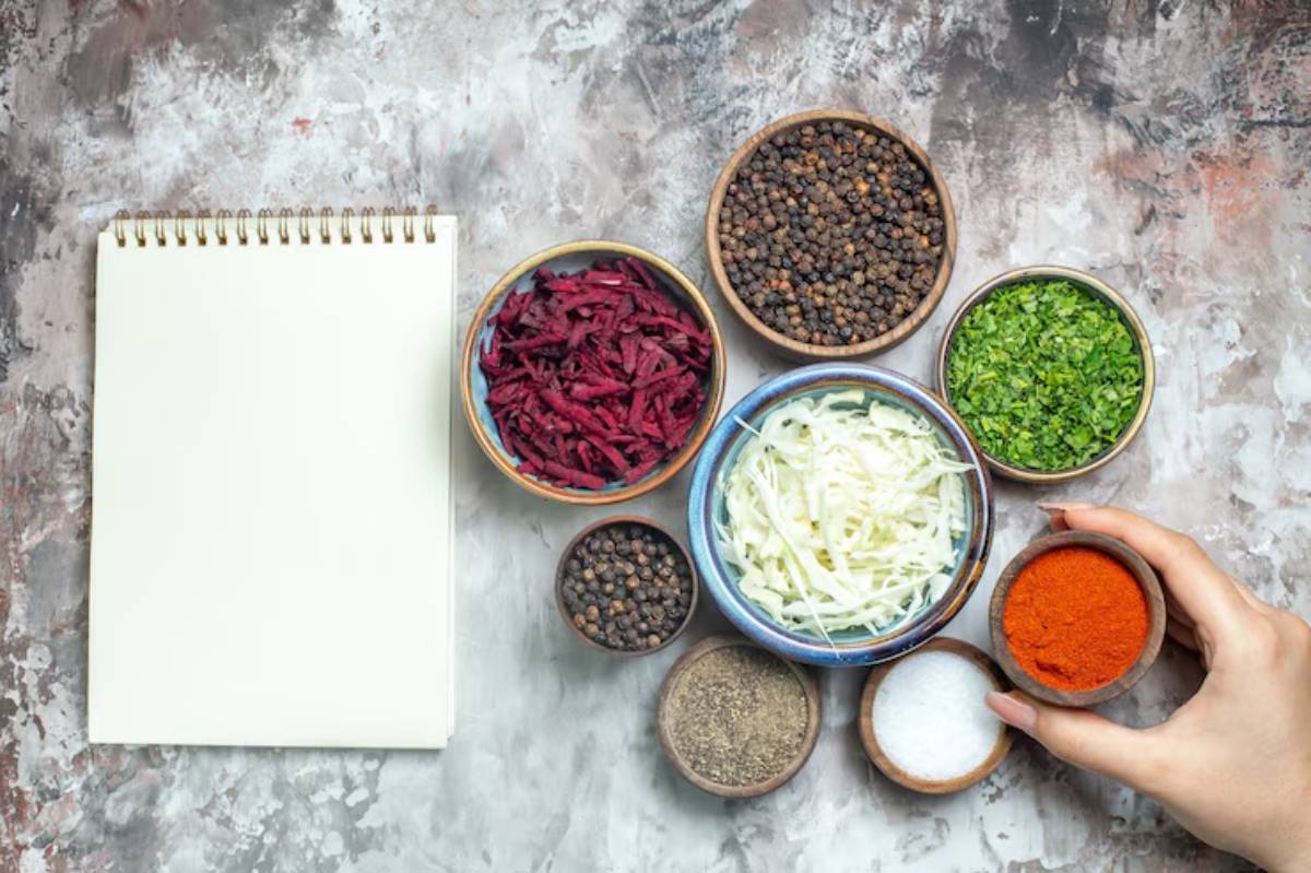 A hand reaches for a bowl of red spice among various spices and vegetables, with a blank notepad nearby on a textured surface.