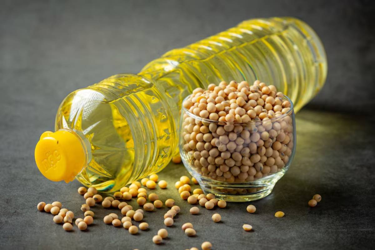 A bottle of yellow cooking oil lies beside a glass bowl filled with soybeans, surrounded by scattered soybean seeds on a dark surface.