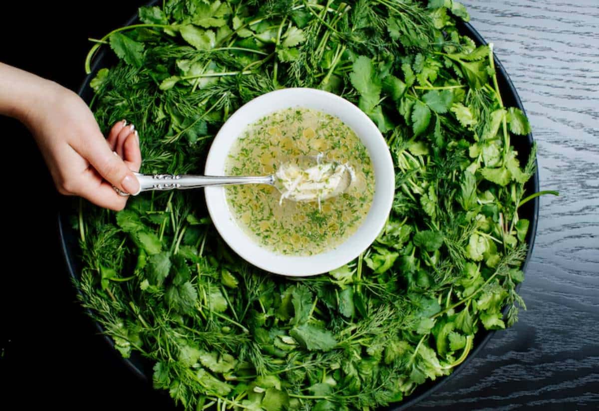 A hand holding a spoon dips into a bowl of broth, surrounded by an array of fresh greens, including cilantro and dill.