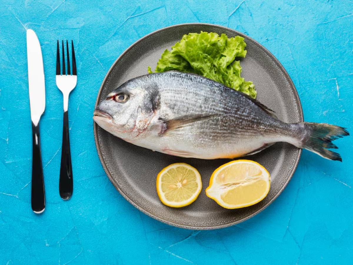A whole fish on a plate with lettuce and lemon slices, accompanied by a knife and fork, set against a bright blue background.