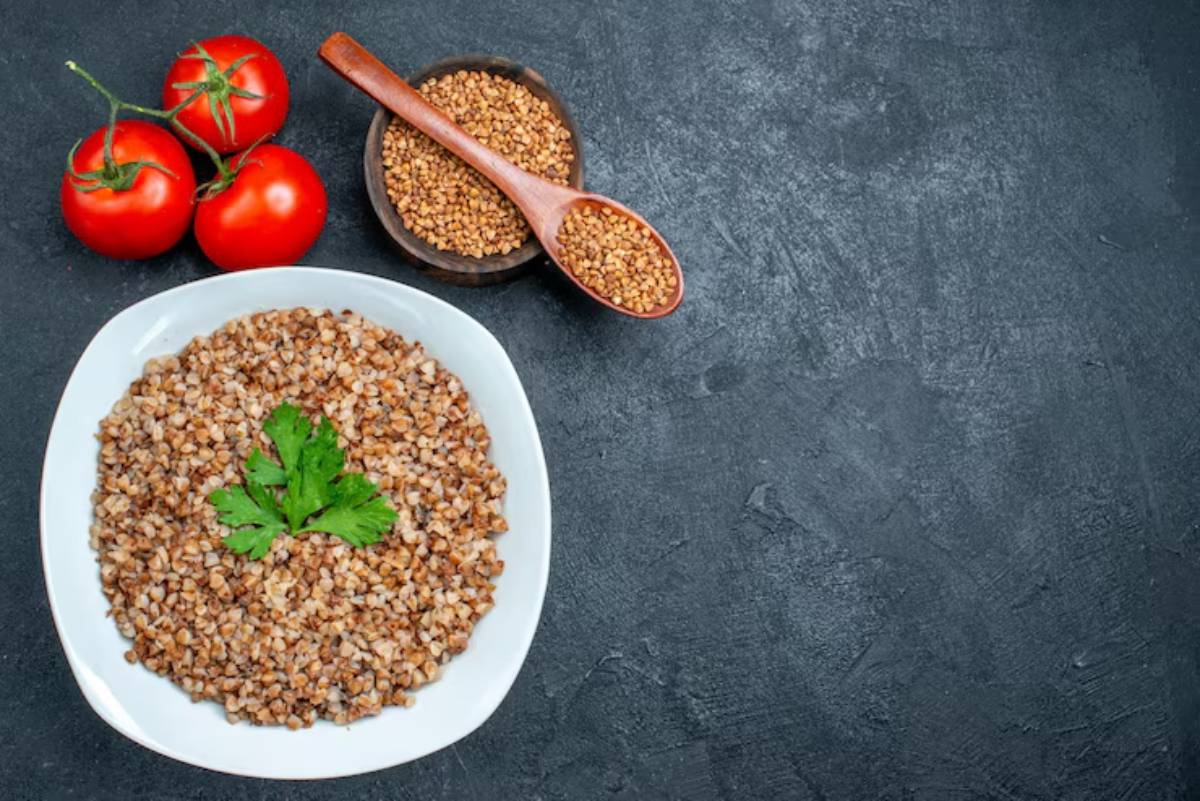 A plate of cooked buckwheat topped with parsley, alongside fresh tomatoes and a bowl of buckwheat groats on a dark background.