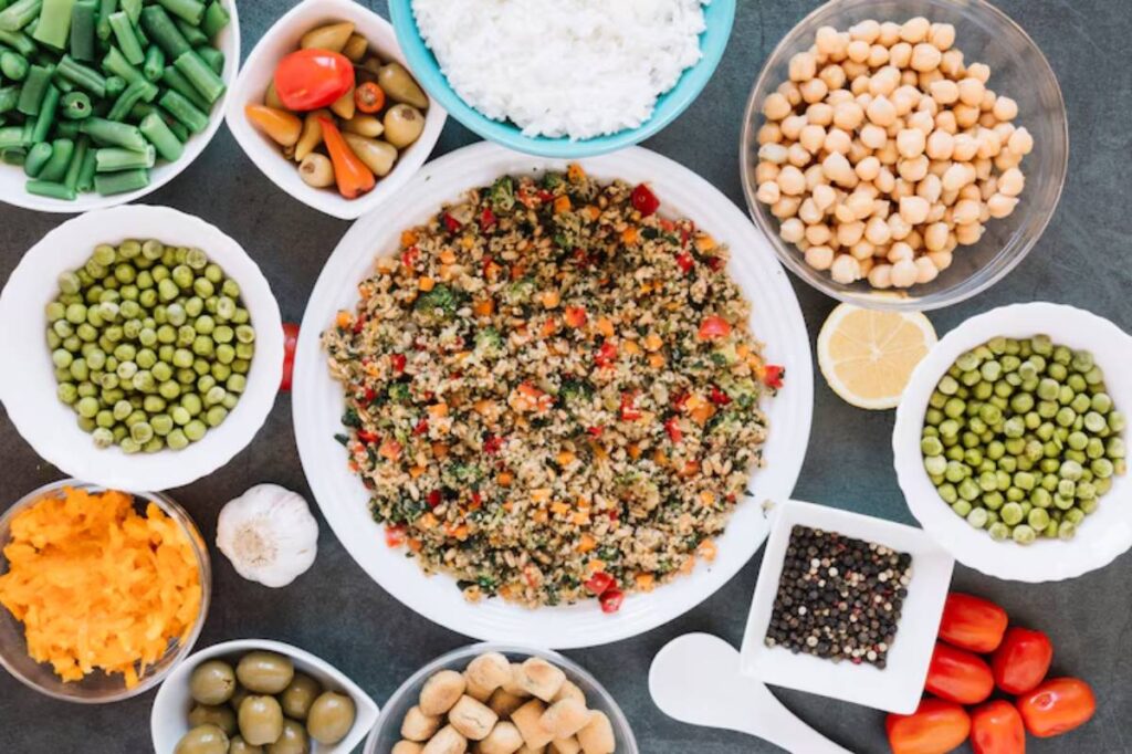 A colorful spread of fresh vegetables, chickpeas, rice, and a quinoa salad, arranged on a dark surface with a lemon slice.