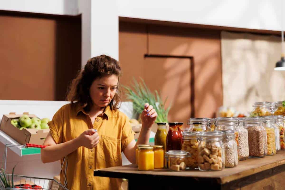 A woman in a yellow shirt inspects products at a market stall filled with jars of various foods, surrounded by fresh produce.