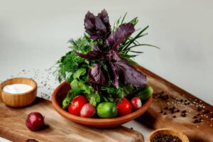 A wooden bowl filled with fresh herbs, tomatoes, cucumbers, and radishes, accompanied by a small bowl of yogurt and spices.